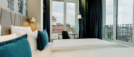 A hotel room with a large bed, a desk, a chair, and a view of the city through the windows.