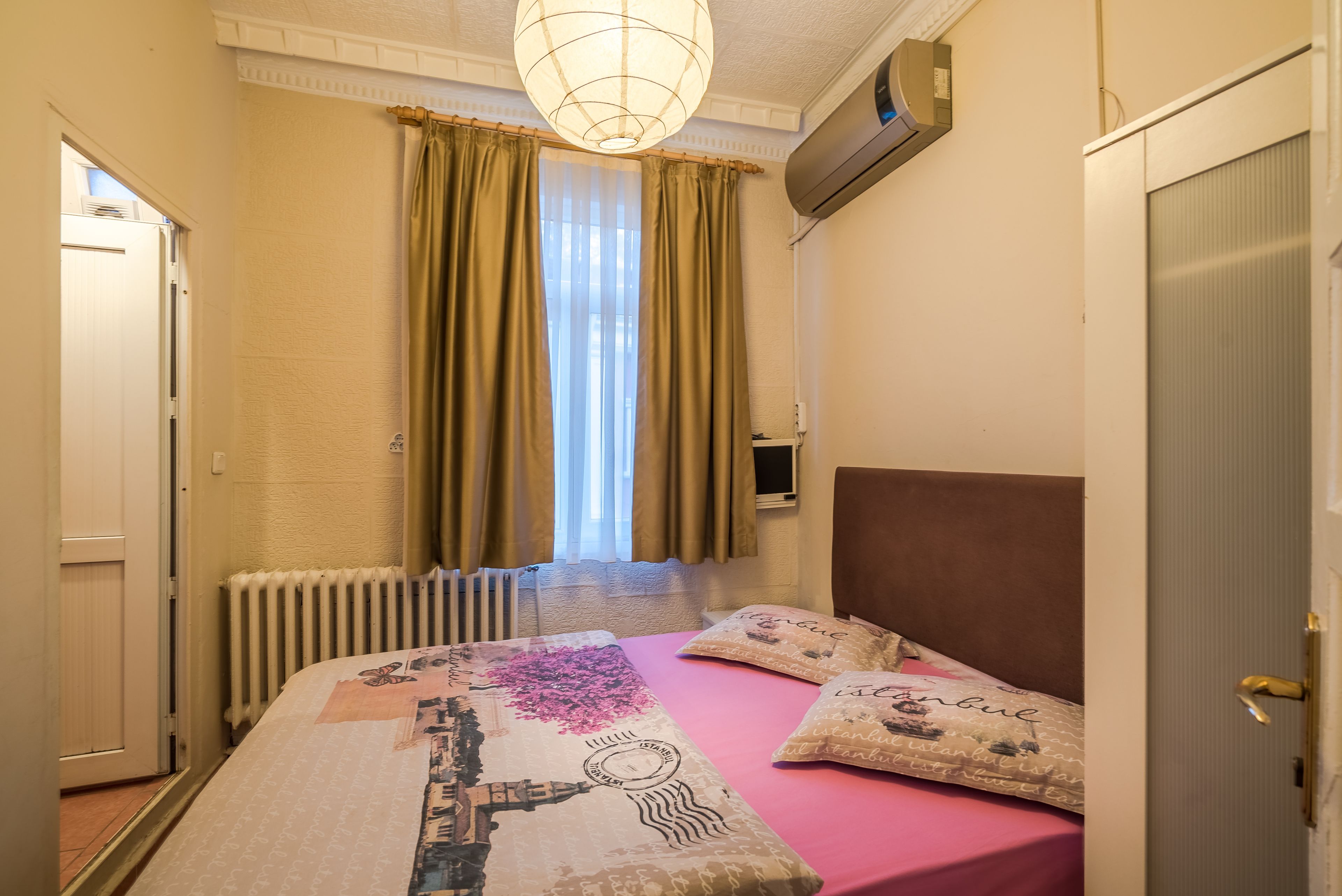 A hotel room with a bed, a radiator, a window with curtains, and a door.