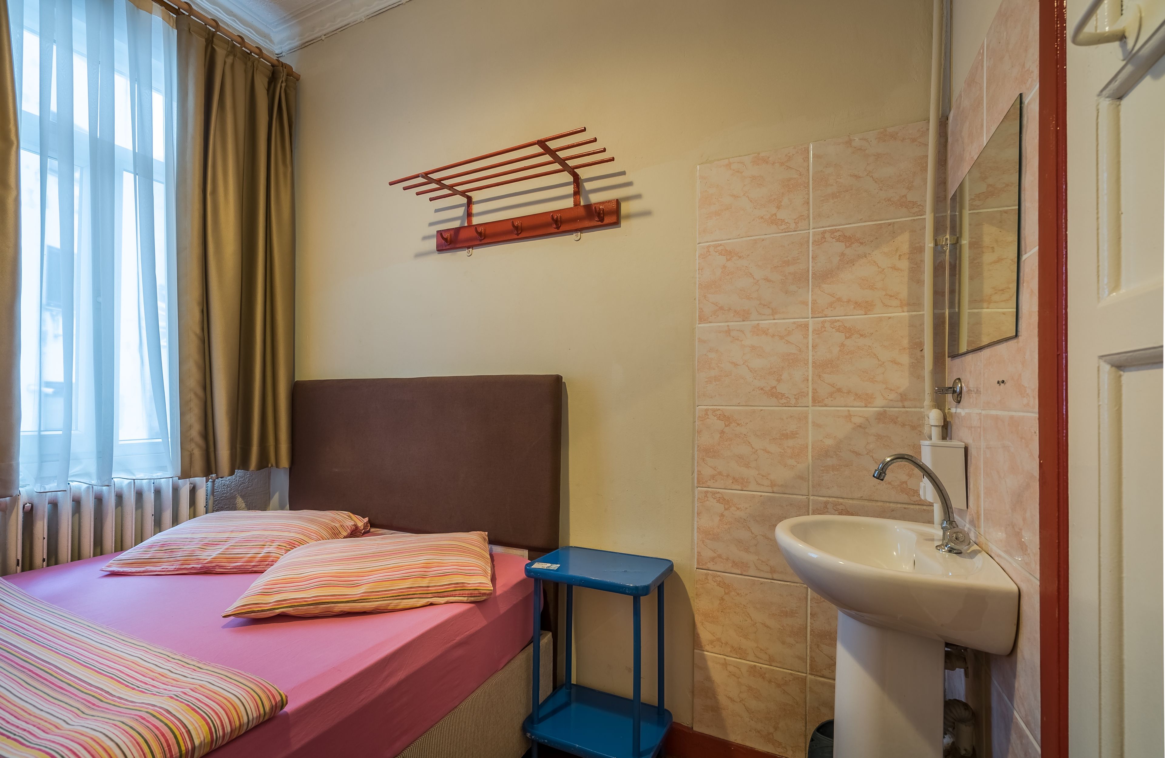 A bedroom with a bed, a blue nightstand, a wall-mounted drying rack, a bathroom with a sink, and a window with curtains.