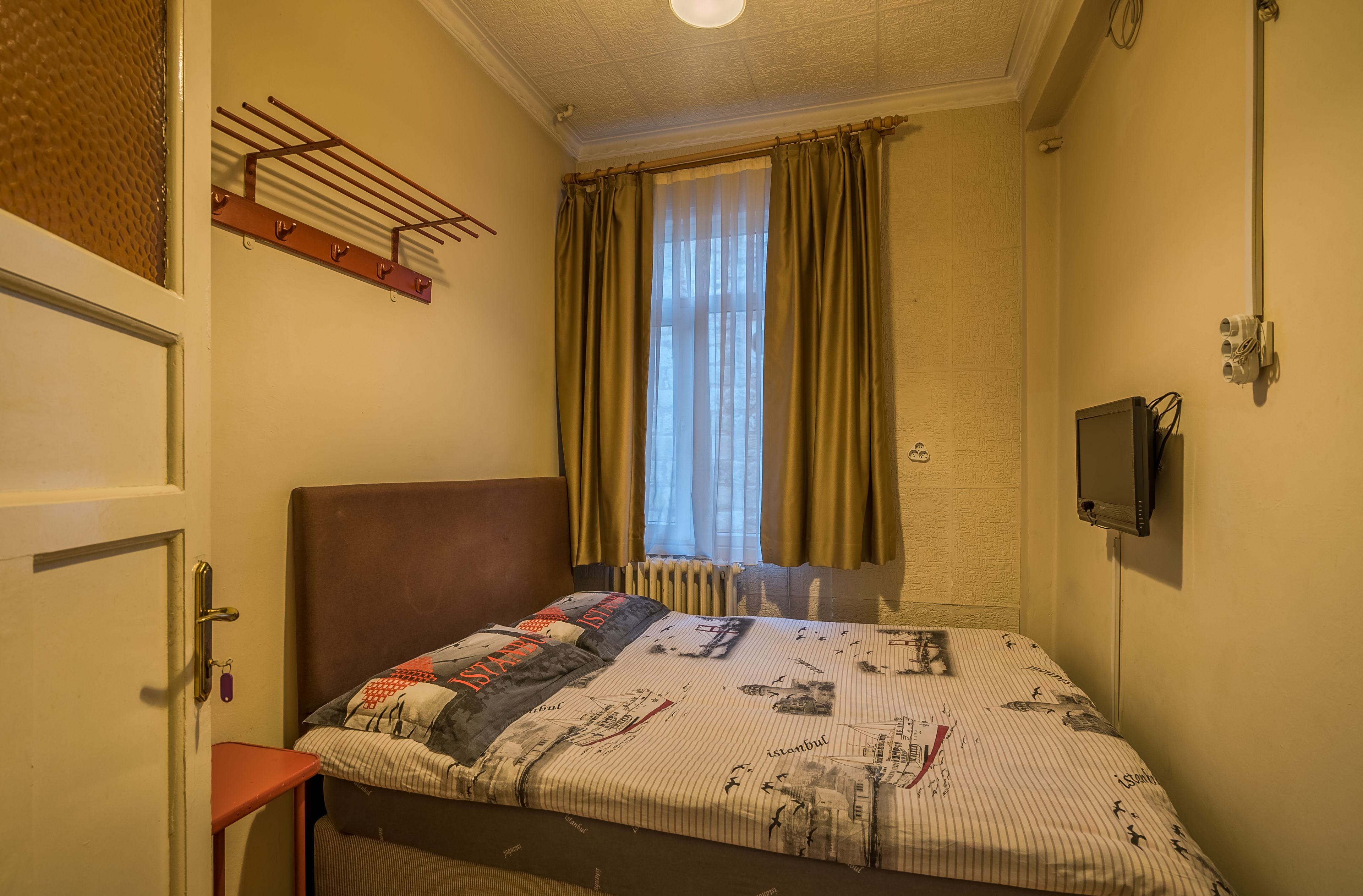 A bedroom with a bed, a window with curtains, a wall-mounted TV, and a radiator.