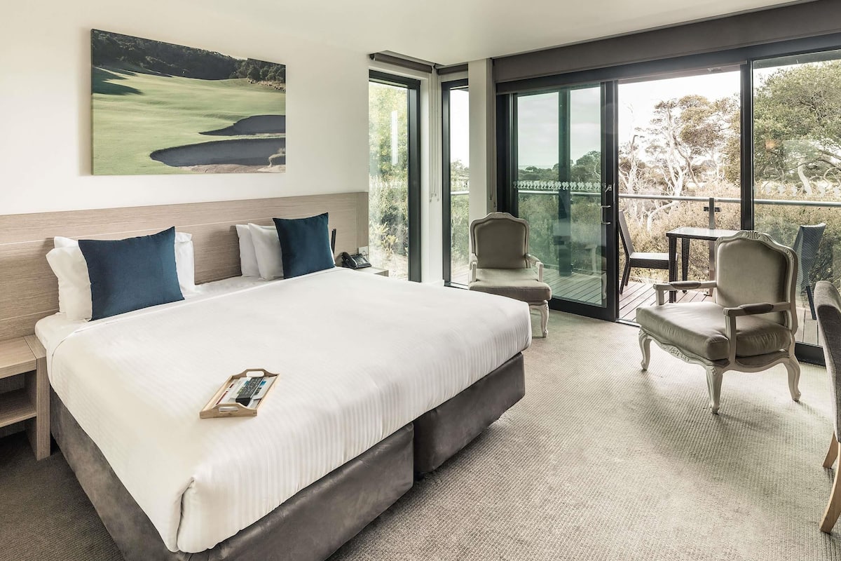 A hotel room with a large bed, a desk, and a chair. There is a window with a view of trees and a golf course.