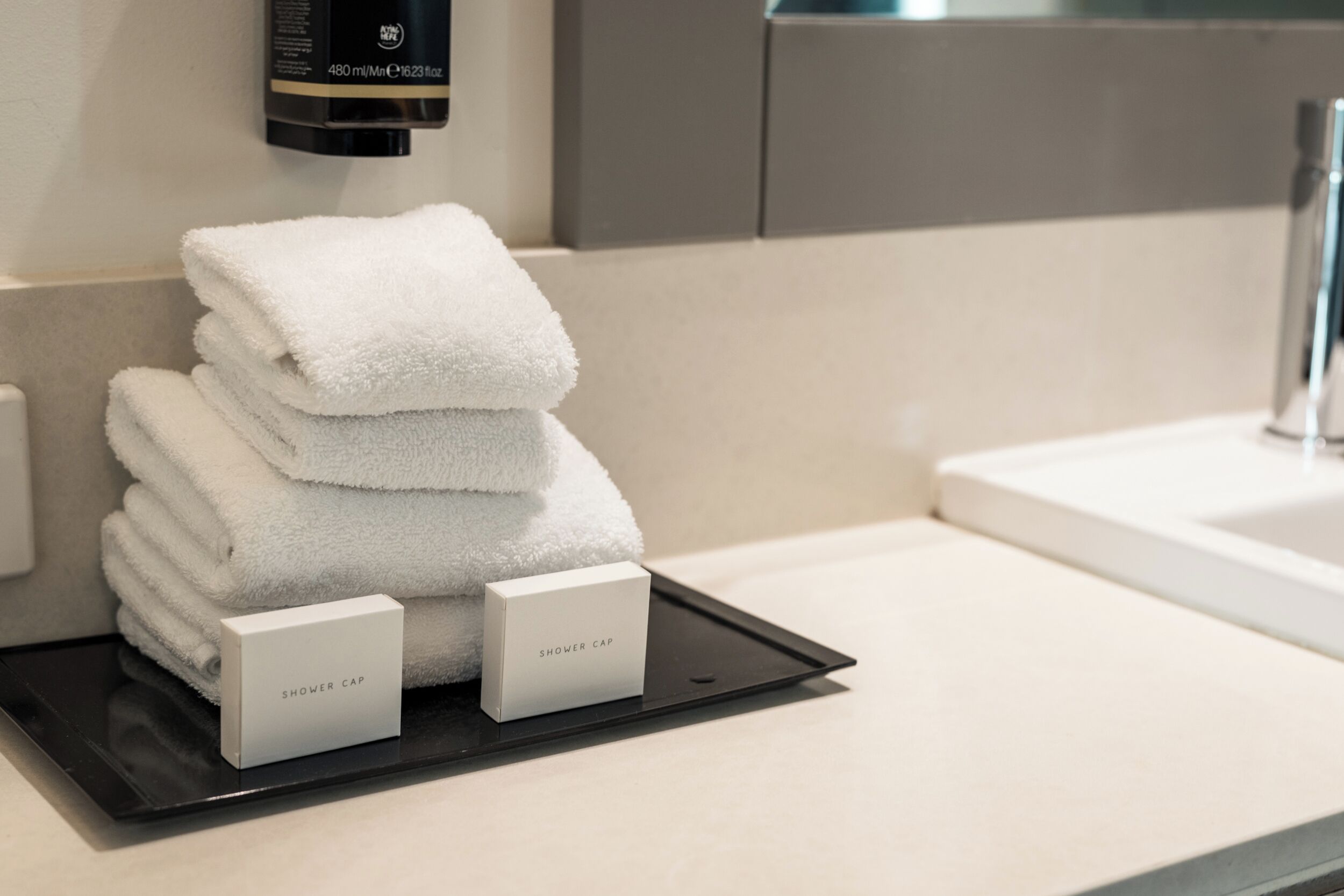 A stack of white towels on a tray with two labeled boxes, a black bottle, and a modern bathroom sink.