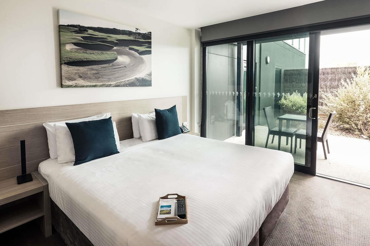 A modern hotel room with a large bed, a bedside table, a nightstand, a glass door leading to an outdoor area, and a painting on the wall.