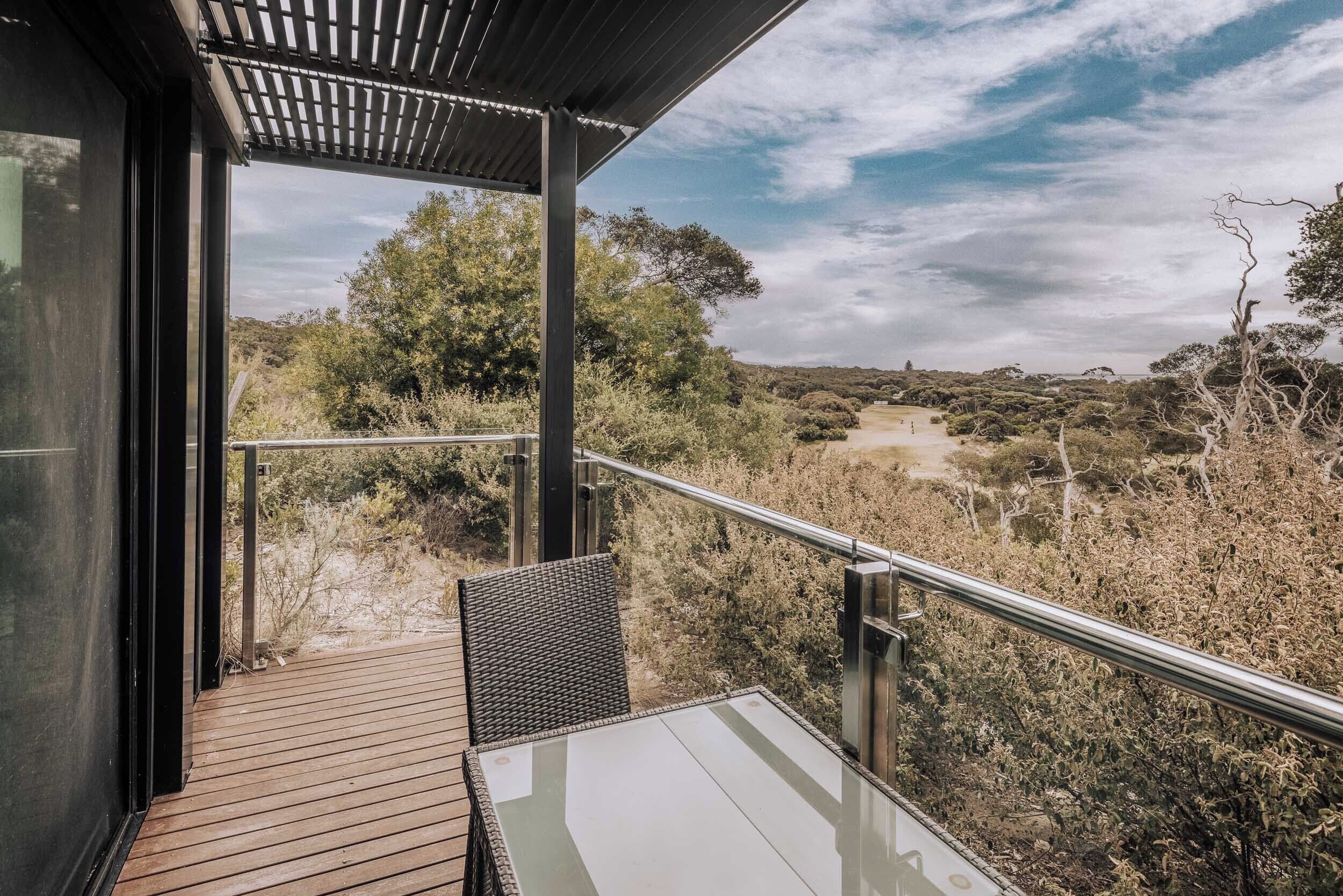 A balcony with a glass table and wicker chair, overlooking a dry landscape with trees and a dirt path.