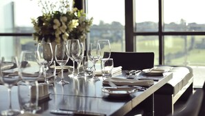 Mesa de comedor dispuesta con copas de vino, platos y cubiertos, con vistas a un paisaje urbano a través de amplios ventanales.