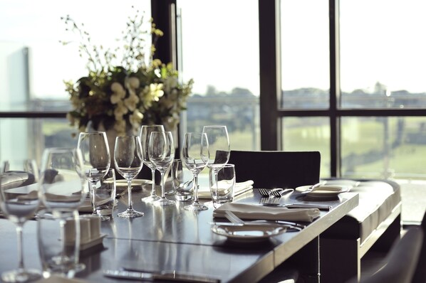 A dining table set with wine glasses, plates, and cutlery, with a view of a cityscape through large windows.
