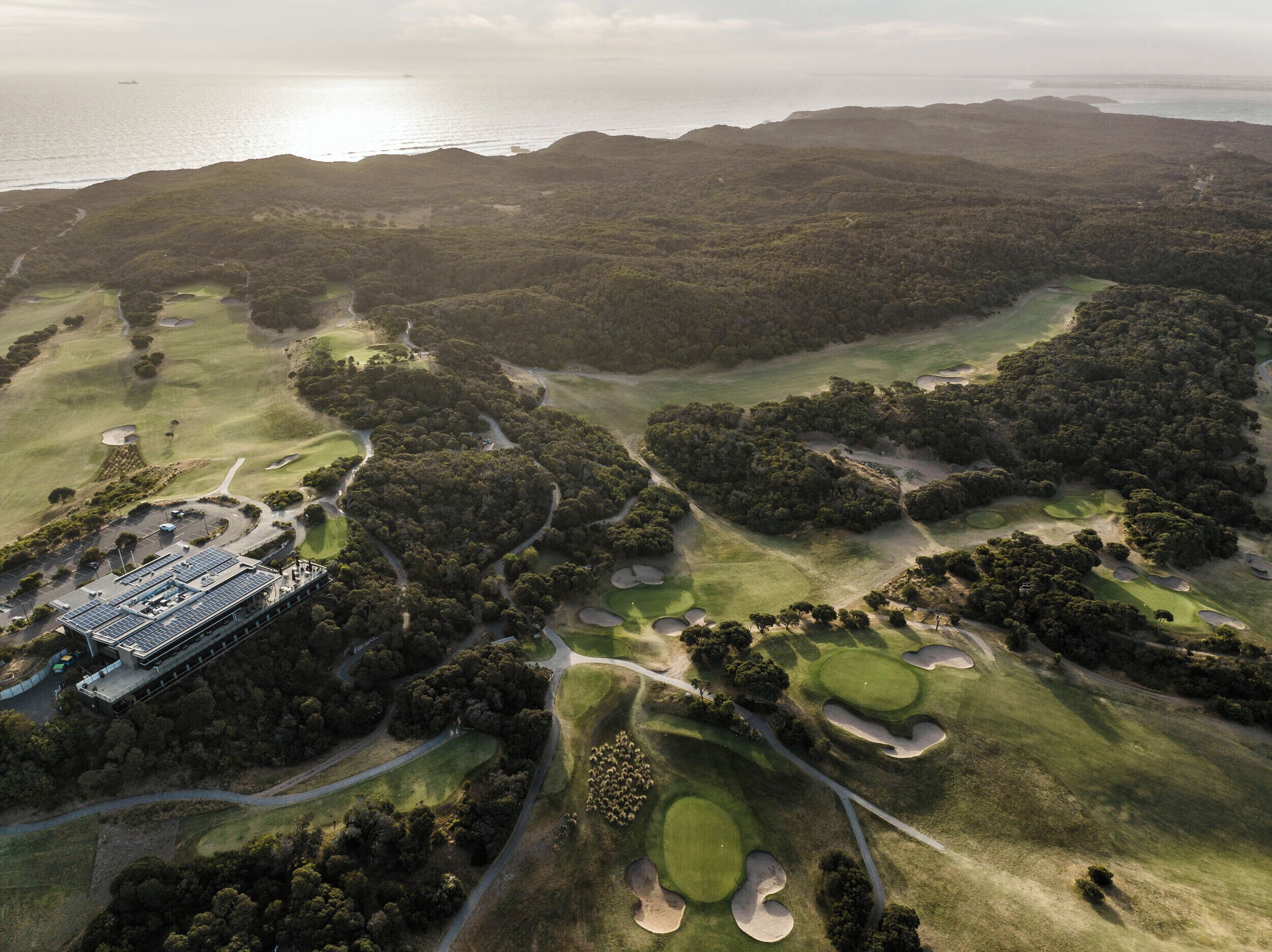 Luchtbeeld van een golfbaan met verschillende holes, een clubhuis en een waterpartij op de achtergrond.