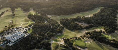 Luchtbeeld van een golfbaan met verschillende holes, een clubhuis en een waterpartij op de achtergrond.