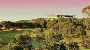 Un edificio moderno con techo plano y amplios ventanales, rodeado de árboles frondosos y un campo de golf.