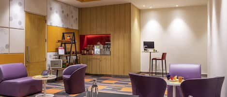 Eine Hotelhalle mit violetten Sitzgelegenheiten, einer Kaffeestation und einem Fernseher.