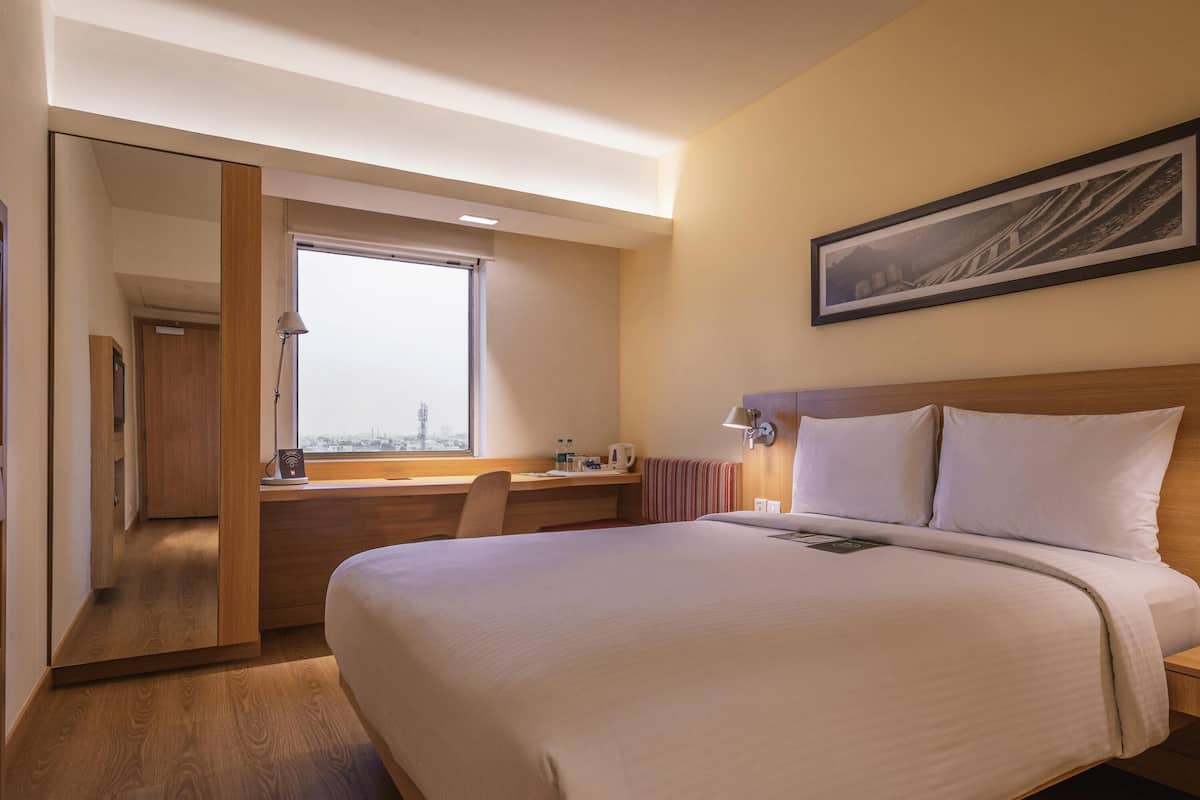 Habitación de hotel con cama, escritorio, silla, ventana con vistas y un cuadro en la pared.