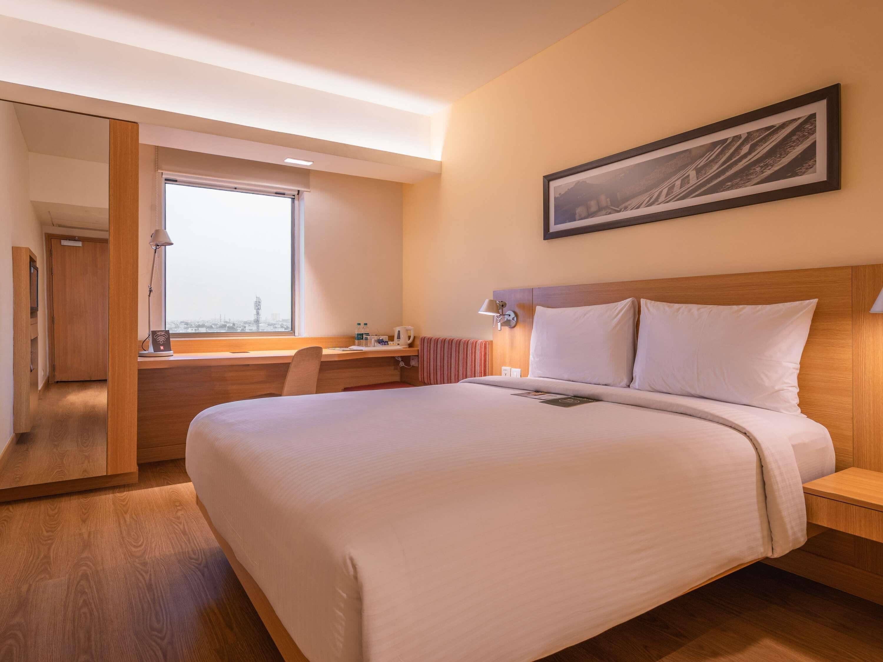 A hotel room with a bed, a desk, a chair, a window, a framed picture, and a door.