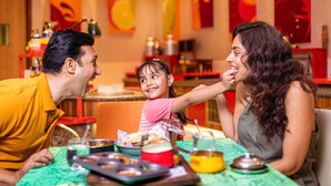 Une famille est assise à une table dans un restaurant, où une nappe verte recouvre le plateau. On peut voir différents plats et des œuvres d’art colorées ornent les murs.