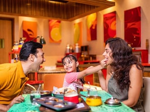 Una familia cenando en un restaurante con mantel verde, diversos platos y coloridas obras de arte en las paredes.