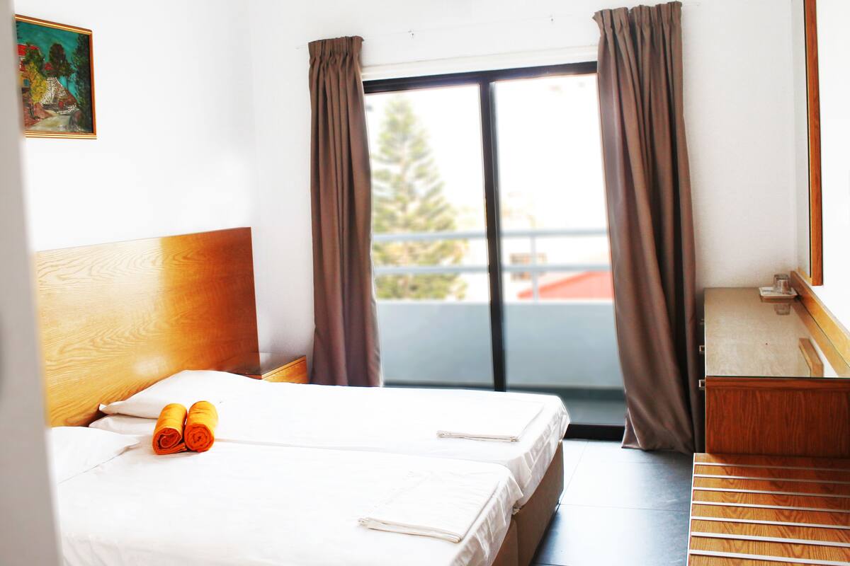 Habitación de hotel con cama, cabecera de madera, escritorio y vistas al exterior a través de una ventana con cortinas.