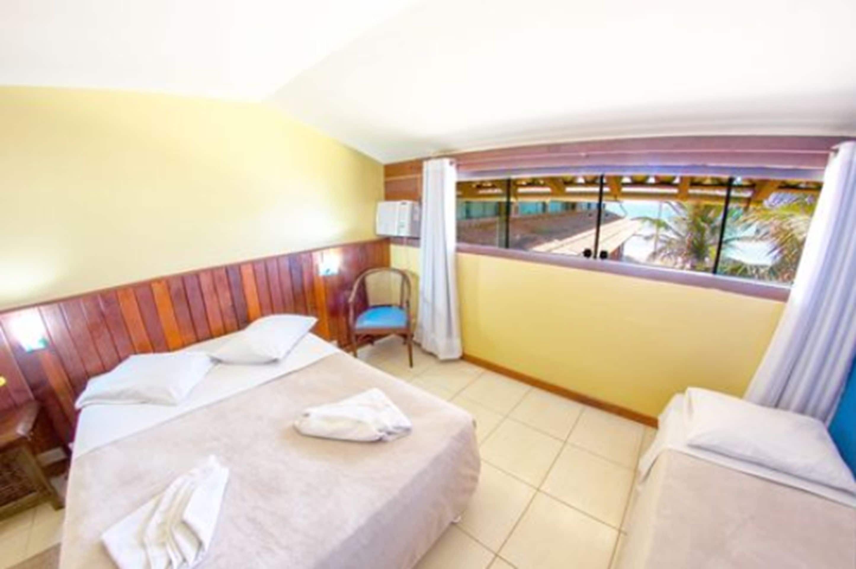 Ein Hotelzimmer mit Bett, Sessel, einem Fenster mit Vorhängen und Blick auf Palmen.