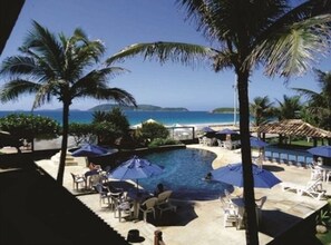 A pool area with lounge chairs and umbrellas, surrounded by palm trees and a view of the ocean. - Hotel La Plage (Cabo Frio)