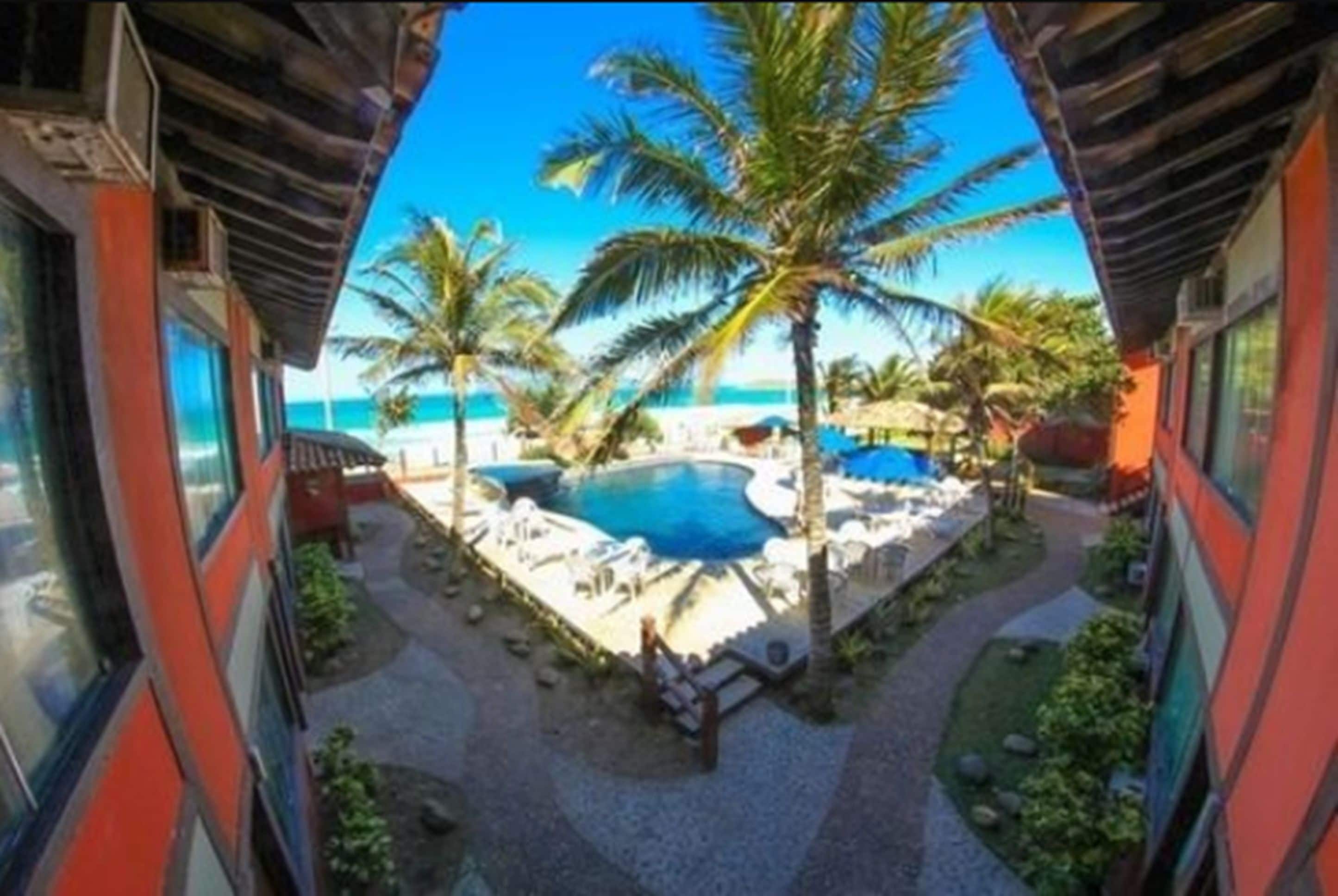 Un área de piscina con palmeras y sillas de descanso, vista al océano, y un edificio con paredes exteriores rojo y blanco.