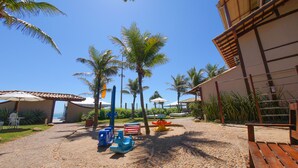 Un sendero que conduce a un edificio de techo con tejas, rodeado de palmeras y un parque infantil con juegos de colores.