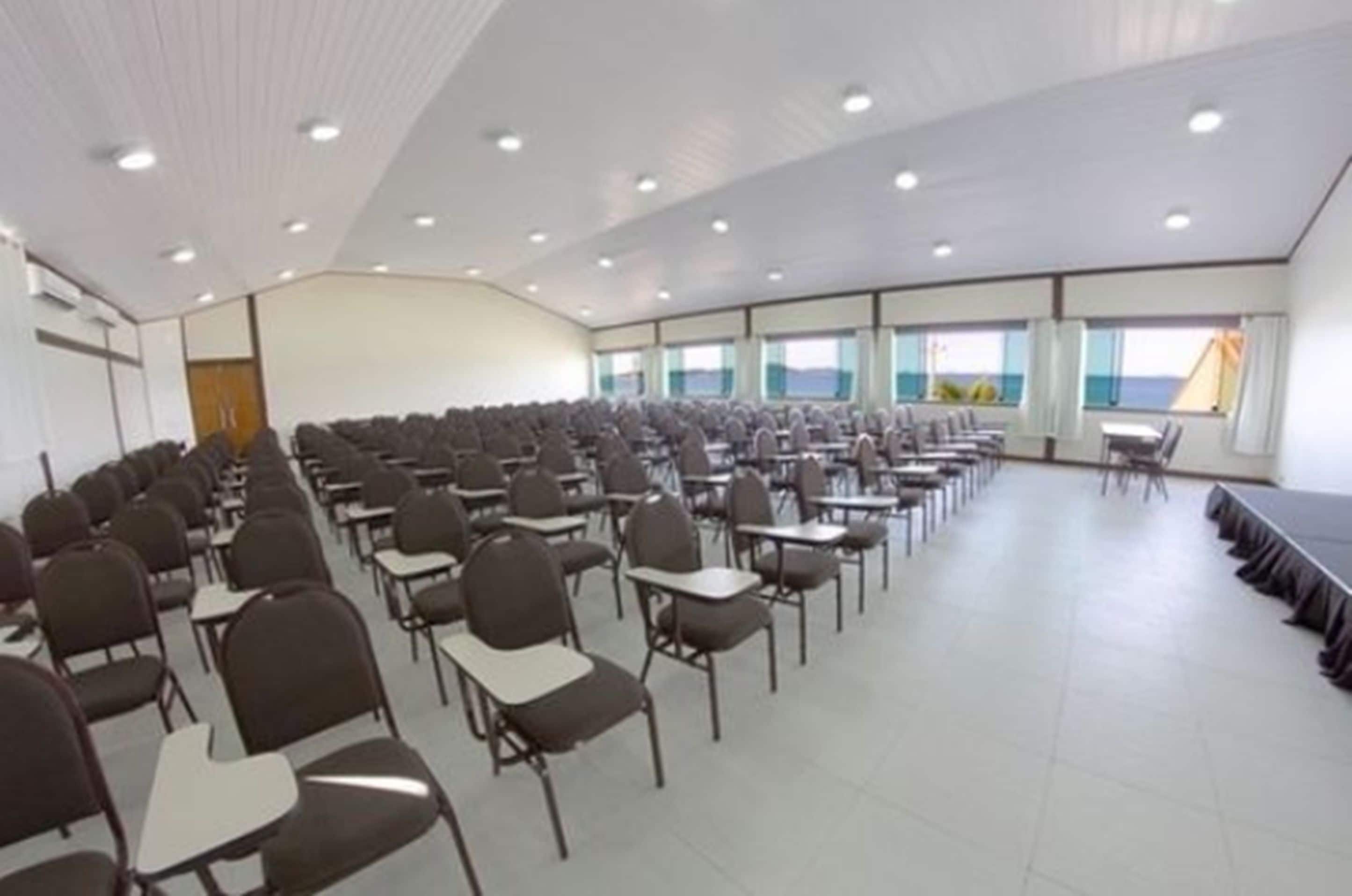 Un salón de conferencias con filas de sillas y mesas, amplias ventanas con vista al mar y un techo blanco con iluminación empotrada.