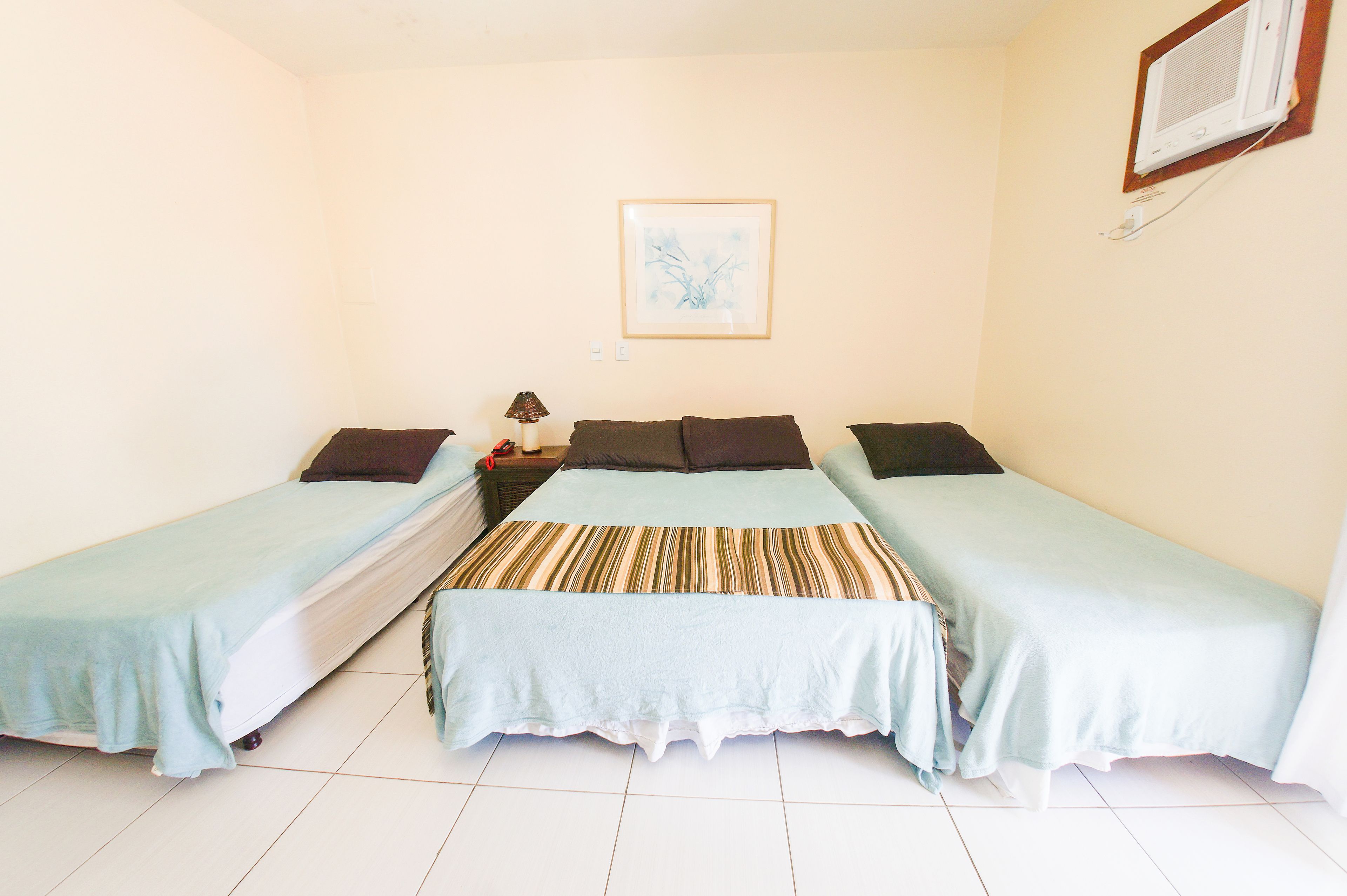 A room with two single beds, a nightstand with a lamp, a framed picture on the wall, and an air conditioning unit.
