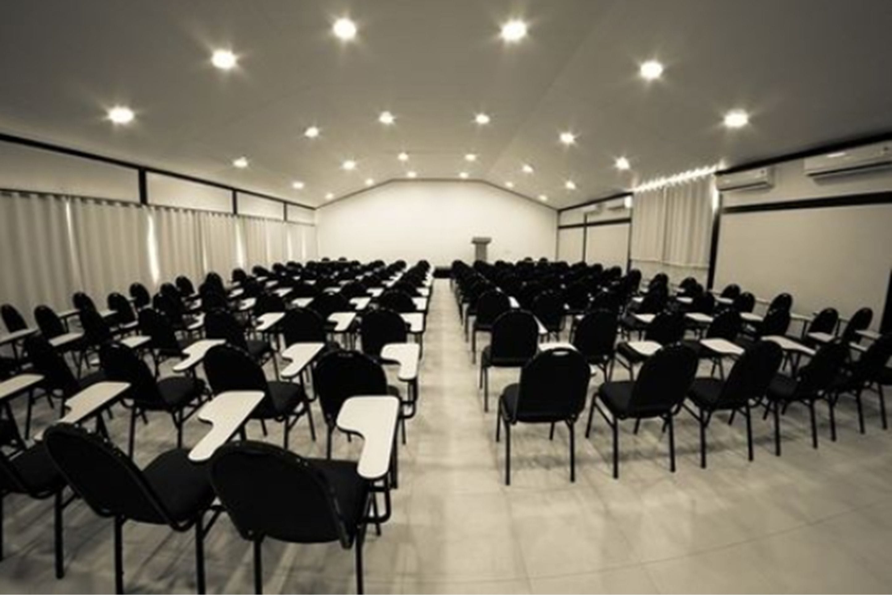 Un salón de conferencias con filas de sillas y mesas, un atril al frente y luces en el techo.
