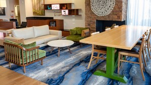 A modern hotel lobby with a wooden dining table, a white sofa, and a green armchair. There is a fireplace with a decorative wall piece above it.
