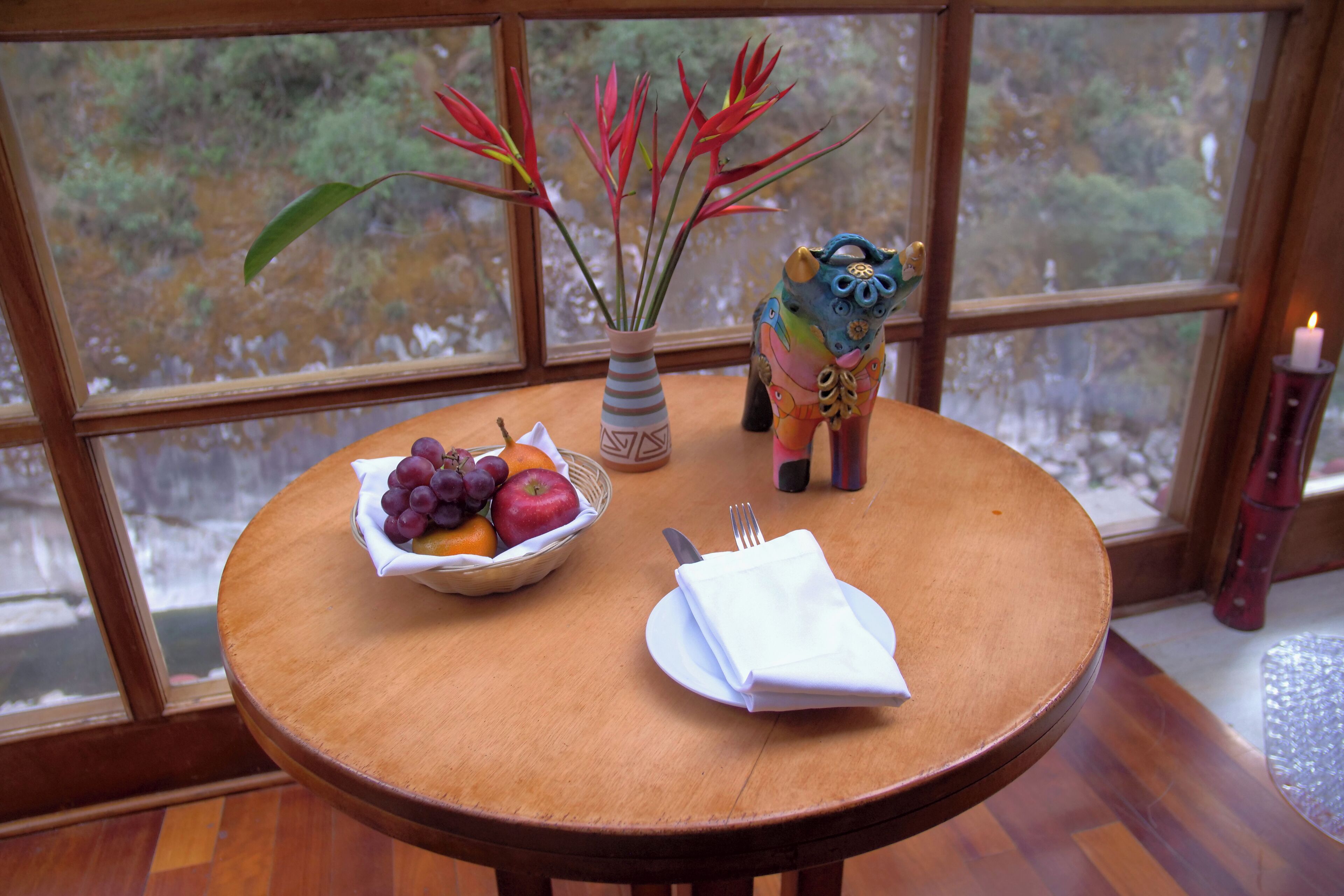 Une table ronde en bois, sur laquelle se trouvent un panier de fruits, un vase avec des fleurs rouges et une statuette de hibou en céramique.