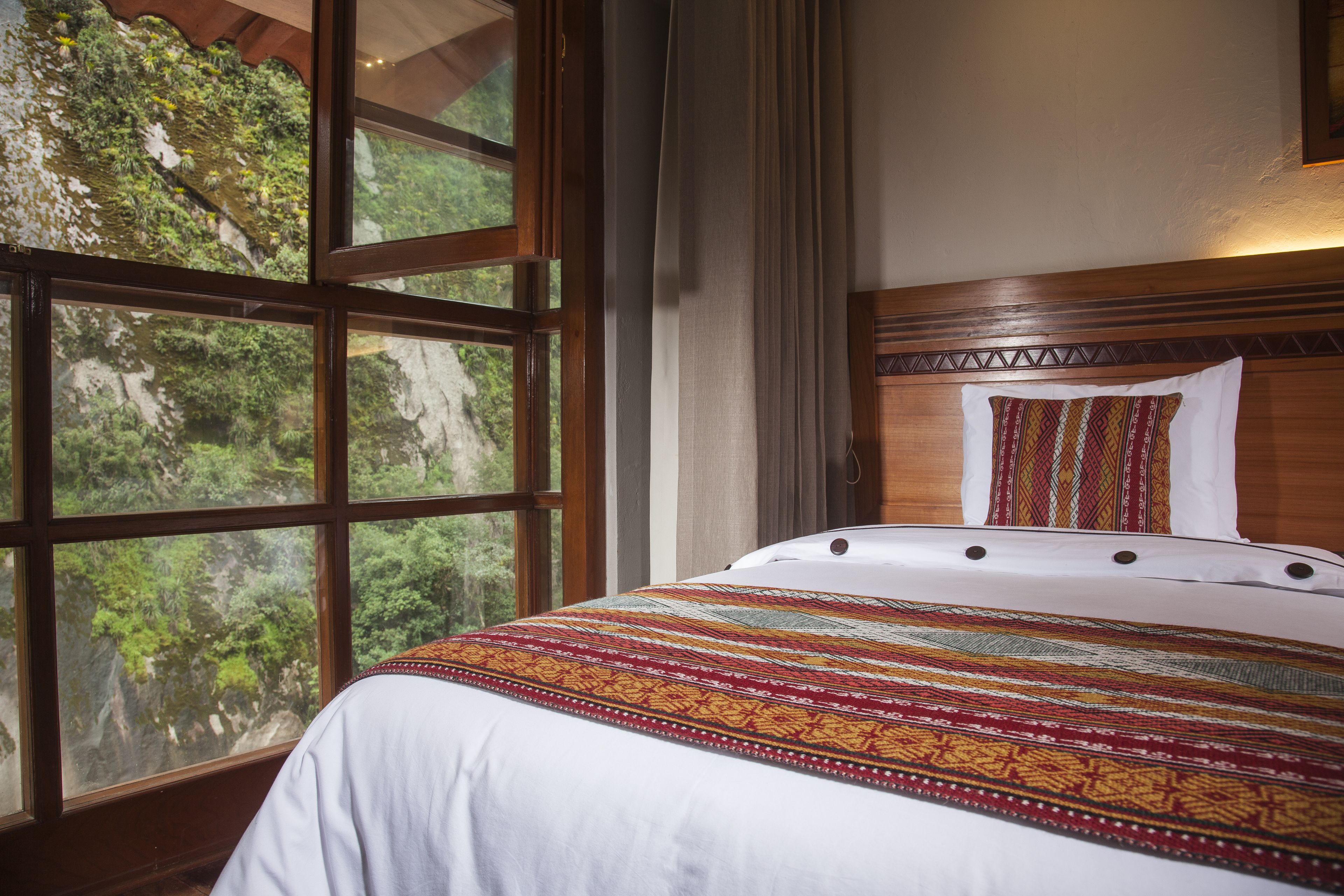 A bed with a patterned bedspread, a wooden headboard, and a window with a view of greenery.