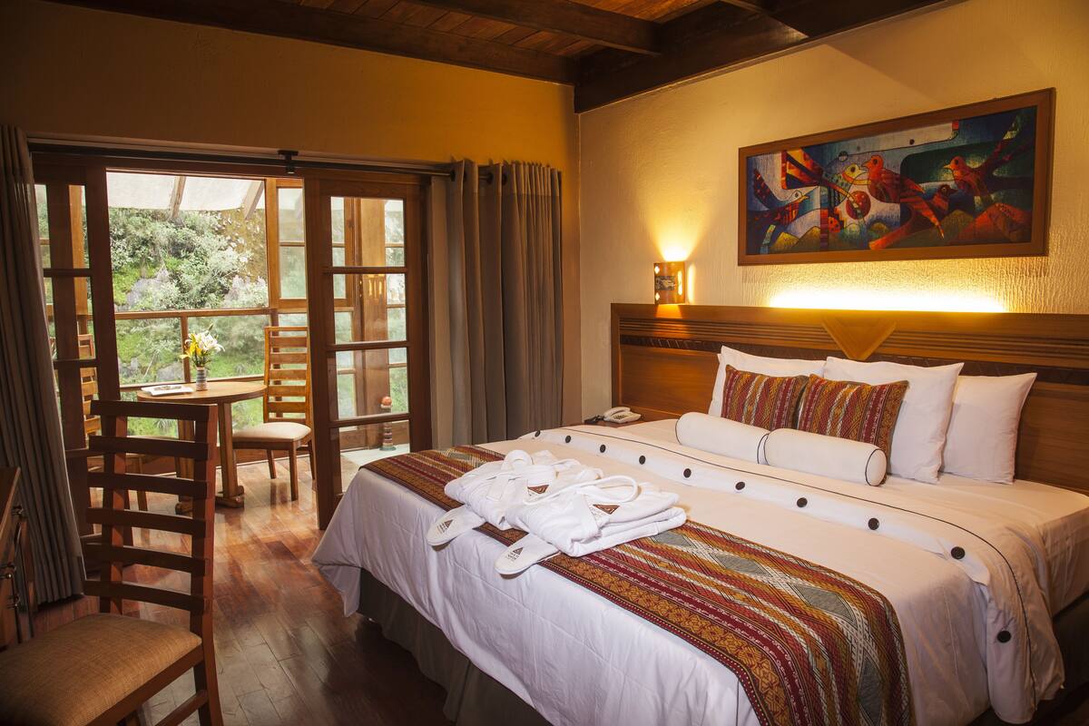 A hotel room with a large bed, a dining table, chairs, a painting on the wall, and a view of the outdoors through the window.