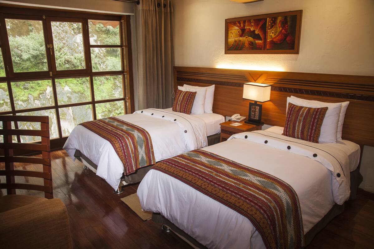 A hotel room with two beds, a wooden chair, a nightstand with a lamp, and a window with curtains.