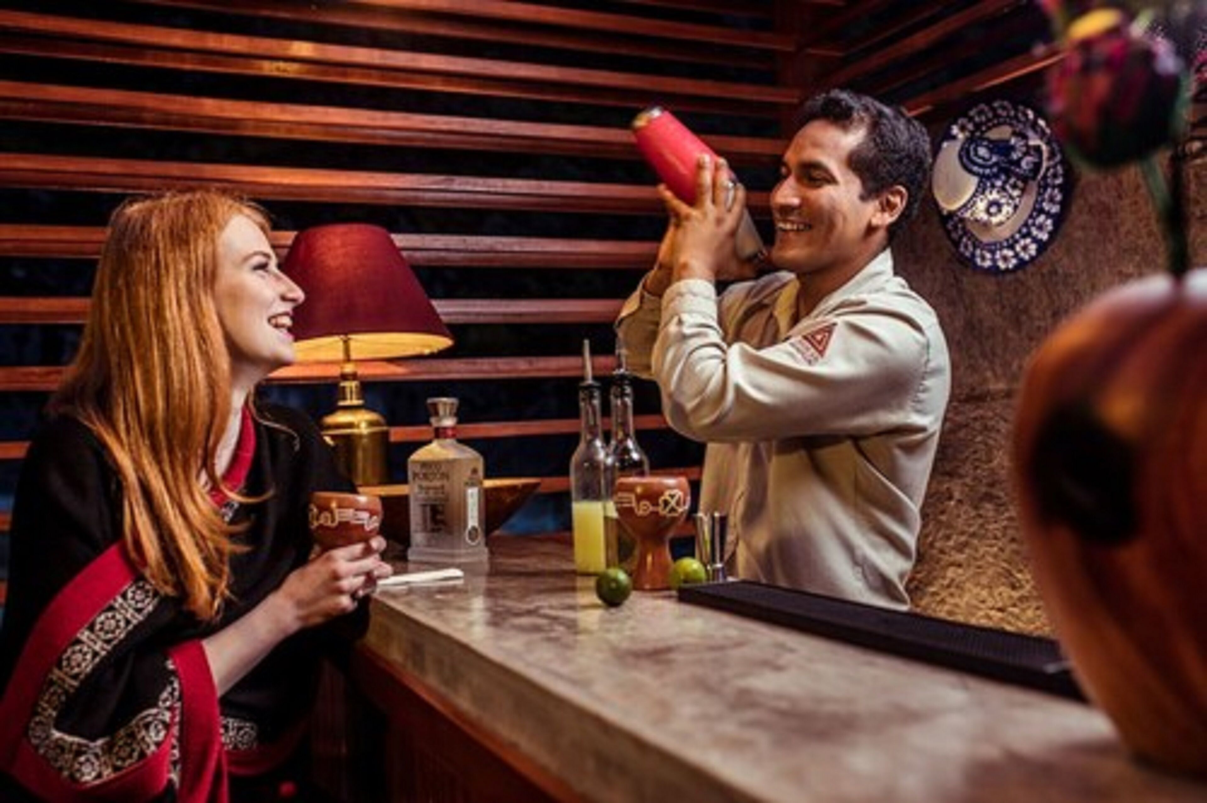 Un bar con un cantinero preparando una bebida, una mujer sosteniendo un vaso, y diferentes utensilios y botellas en la barra.