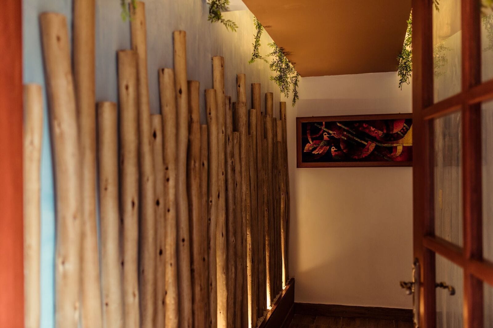 Pared de listones de madera con plantas colgantes, una obra de arte enmarco y una puerta de madera.