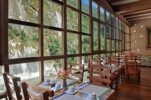 A dining area with large windows, wooden furniture, and a view of a waterfall. - Casa Del Sol Machupicchu (Machu Picchu)