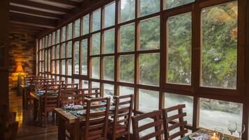 A restaurant with wooden tables and chairs, large windows with a view of a forested area.