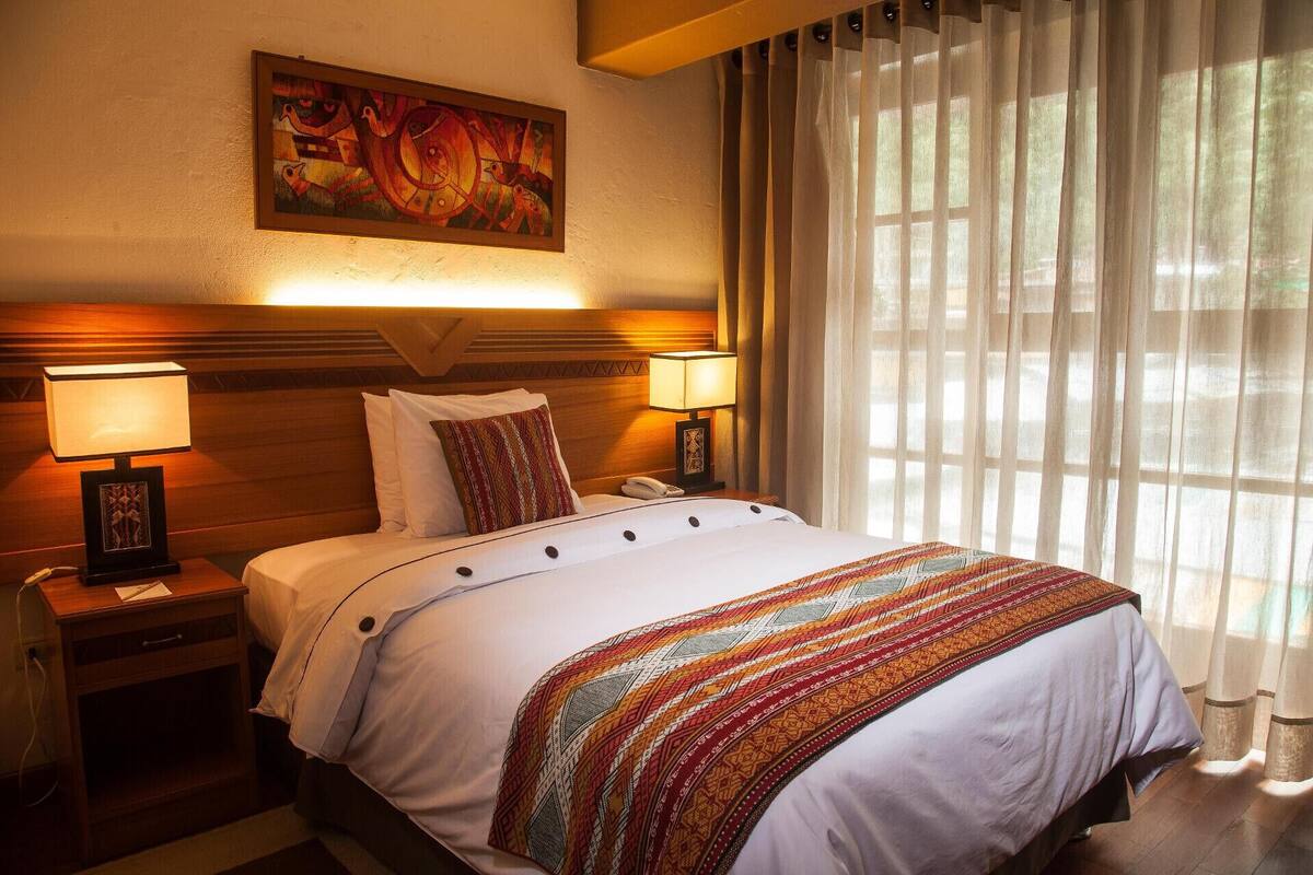 A hotel room with a bed, two bedside lamps, a painting on the wall, and sheer curtains.