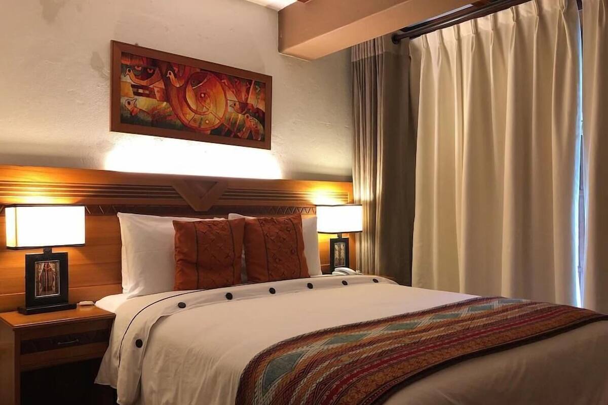 A hotel room with a large bed, two bedside lamps, a headboard, a painting above the bed, and a window with curtains.