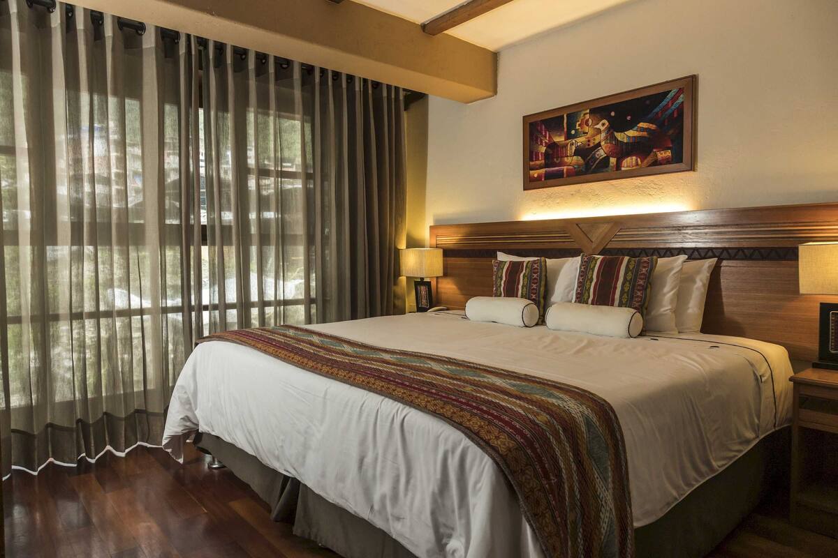 A hotel room with a large bed, a headboard, two bedside lamps, a painting on the wall, and sheer curtains on the window.