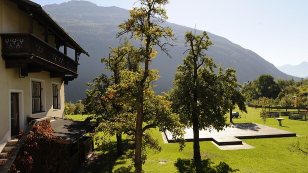 A traditional chalet with a balcony, surrounded by a well-maintained lawn and trees, with mountains in the background.