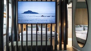 A modern hotel room with a large TV displaying a scenic view, a bed with white linens, and a small seating area with a patterned ottoman.