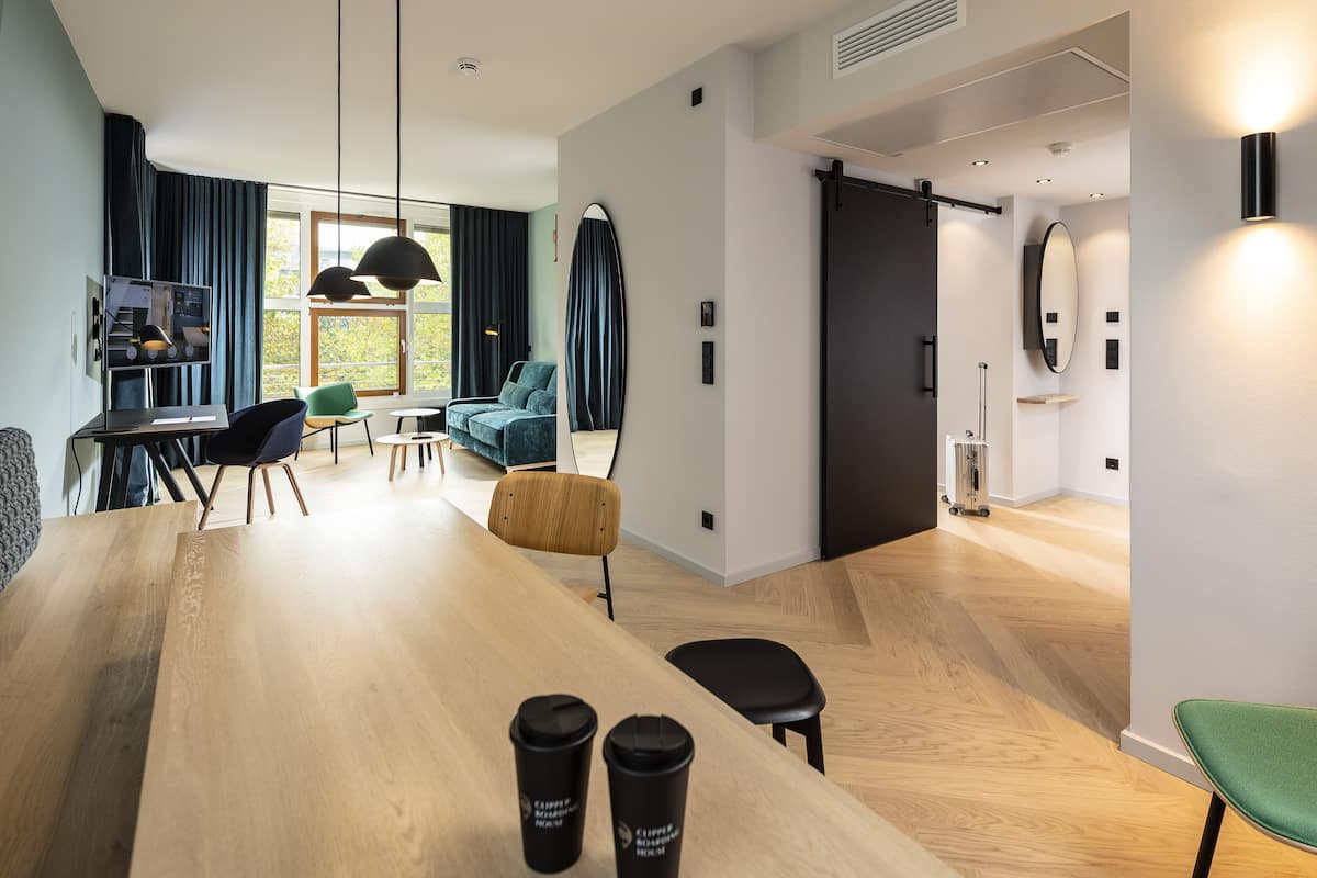 Habitación de hotel moderna con una mesa de comedor de madera, un sofá y una silla verde. Hay dos tazas de café negras sobre la mesa.