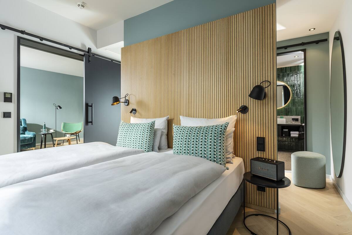 Una habitación de hotel moderna con una cama grande, cabecera de madera con listones, una mesita de noche con lámpara y un otoman verde.