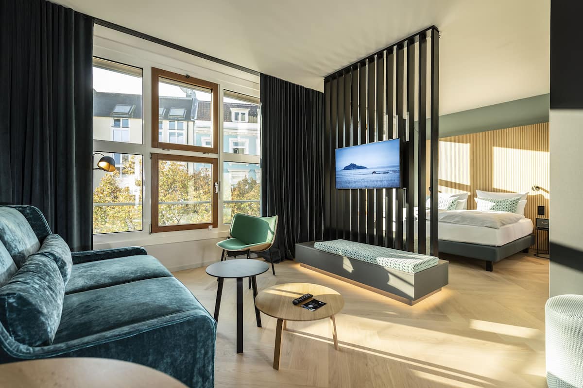 Una habitación de hotel moderna con sofá, una mesita redonda, televisor montado en la pared, cama y ventana con vistas a edificios y árboles.