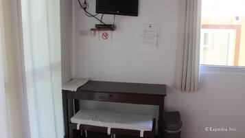 Una habitación con una televisión montada en la pared, un escritorio con una silla, un cesto de basura y una ventana con cortinas.