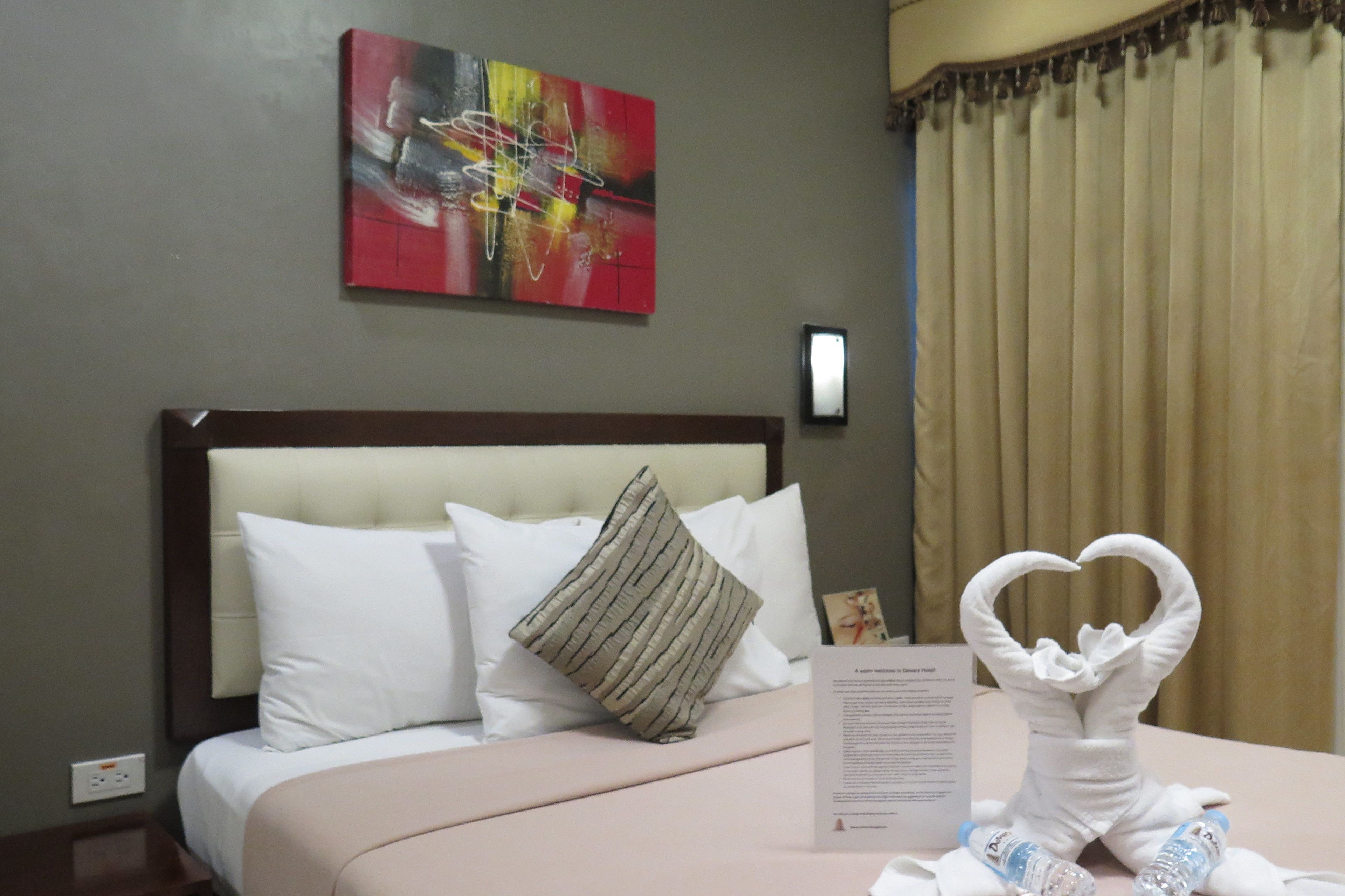A hotel room with a bed, pillows, a heart-shaped towel sculpture, a painting on the wall, and a window with curtains.