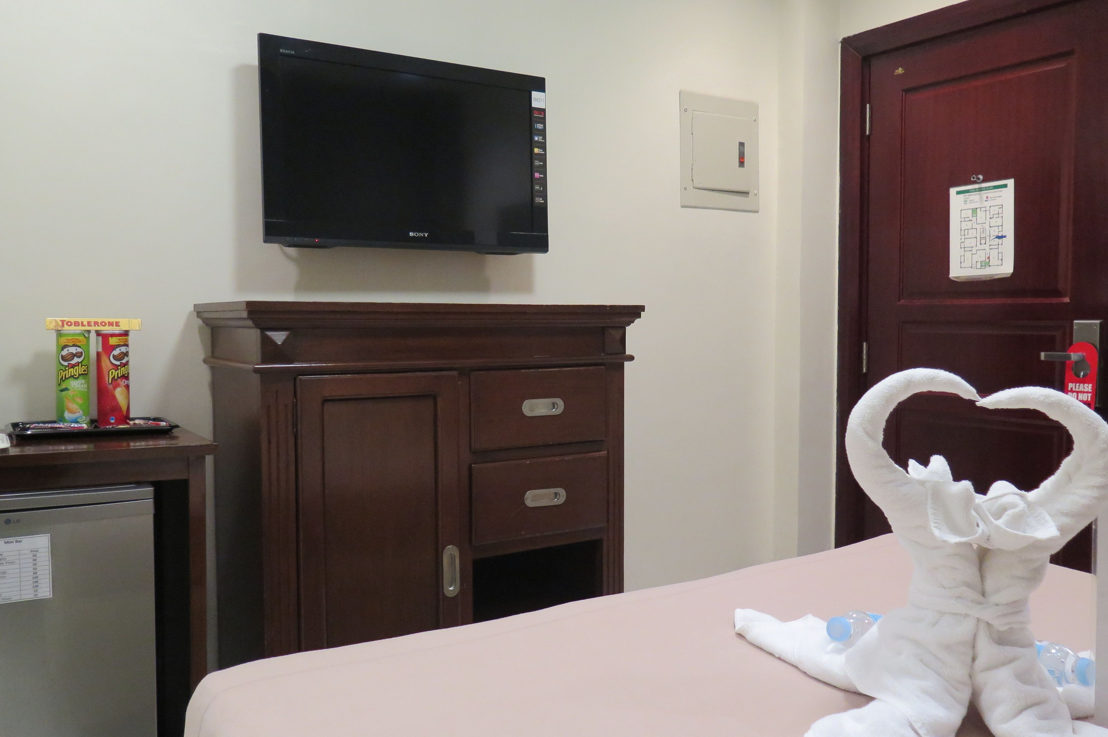 Habitació d'hotel amb televisió de pantalla plana, un armari de fusta, un minibar i una tovallola amb forma de cor sobre el llit.