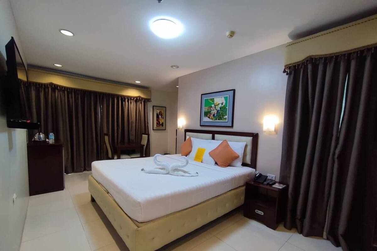 A hotel room with a large bed, a bedside table, a desk, a chair, a flat-screen TV, and a painting on the wall.