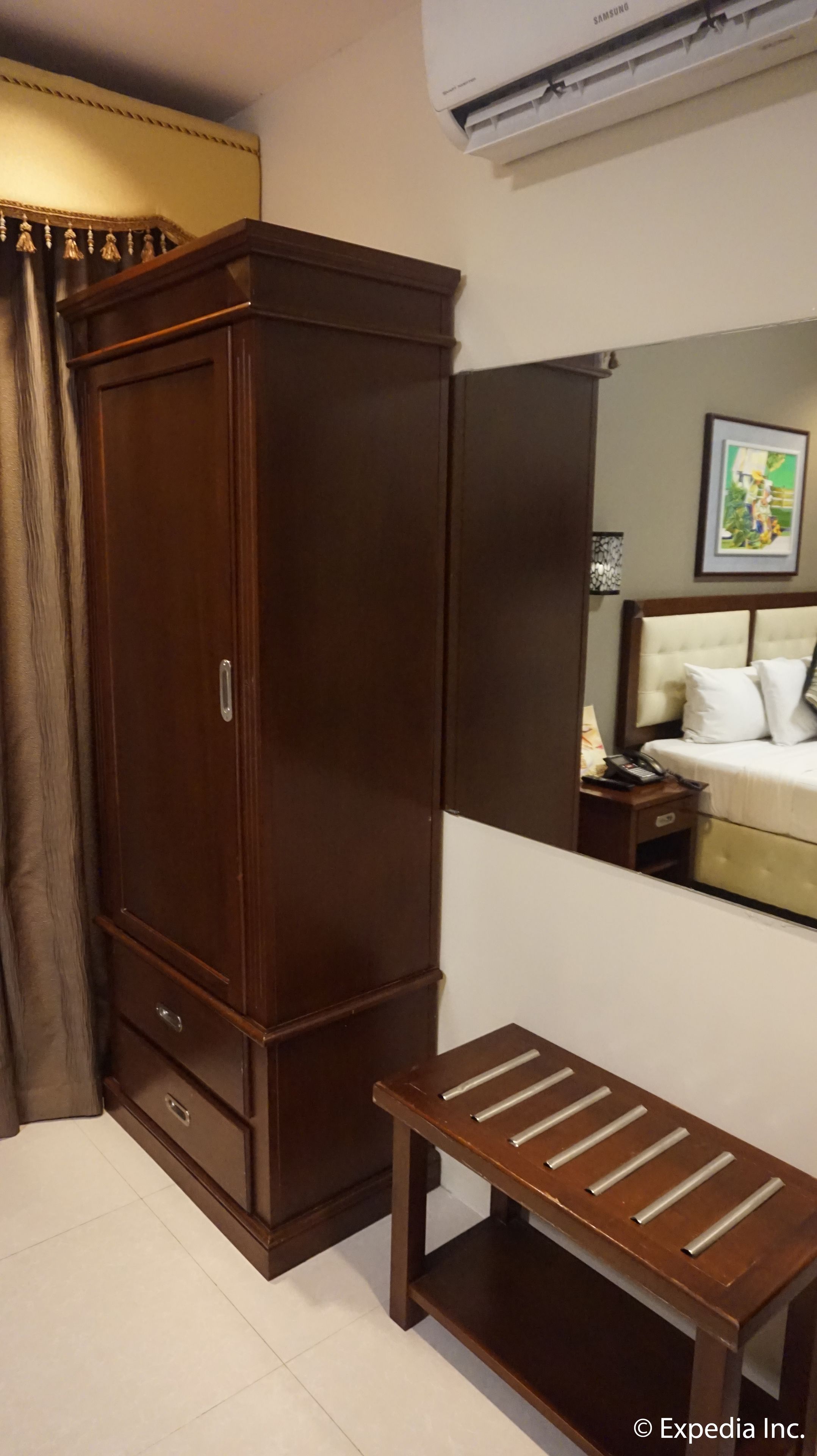 Ein Zimmer mit einem Holzschrank, einer kleinen Holzbank, einem Bett mit weißer Bettwäsche, einem Nachttisch mit einer Lampe und einer wandmontierten Klimaanlage.