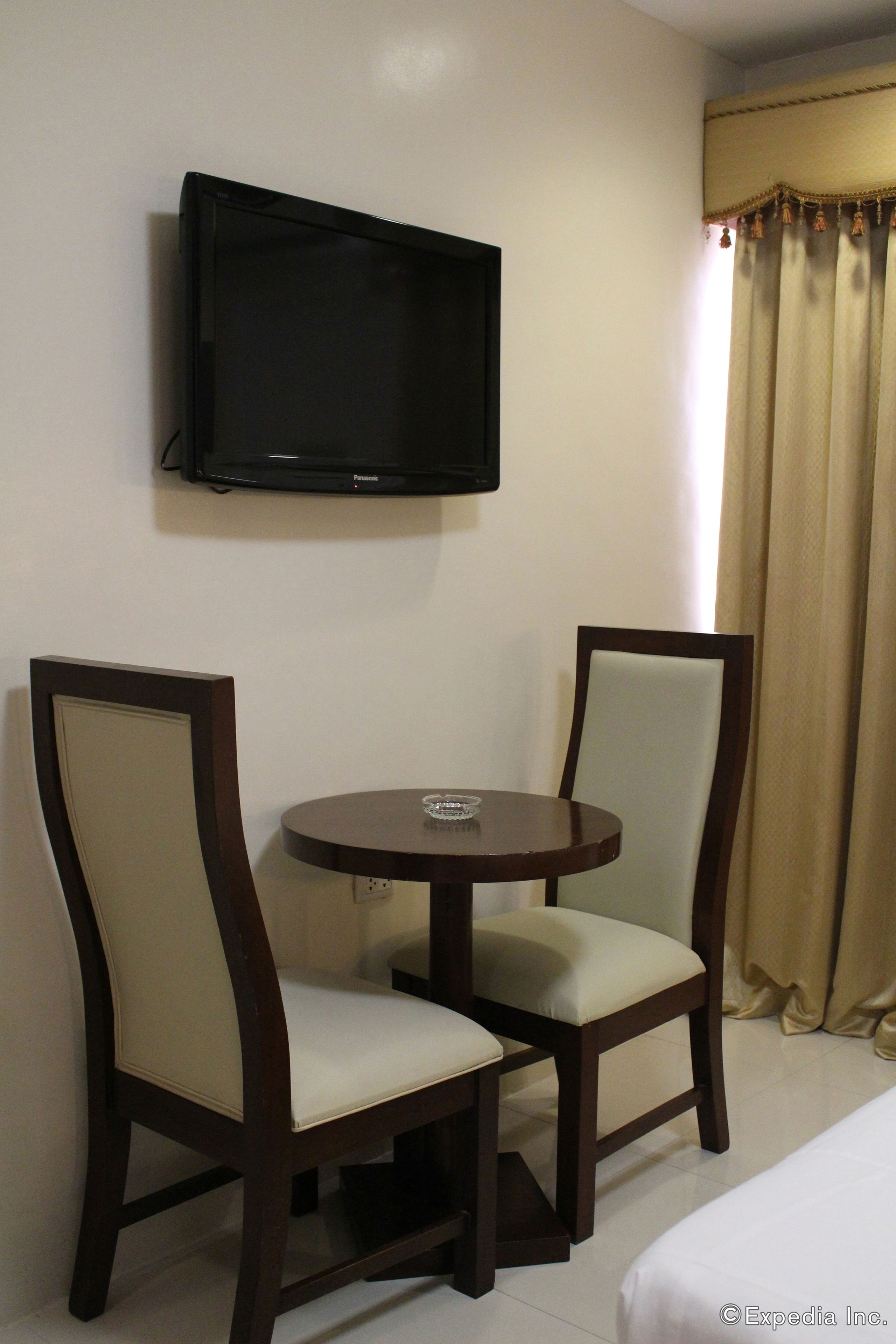 Quarto de hotel com uma cama, uma televisão na parede e duas cadeiras ao redor de uma mesinha.