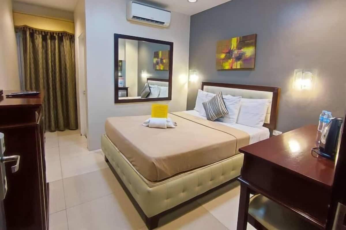 A hotel room with a bed, a desk, a mirror, an air conditioning unit, and a painting on the wall.